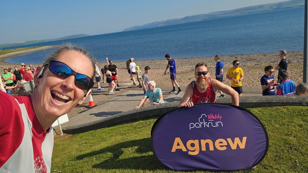 Two runners and the Agnew Parkrun sign with other runners behind and a beach and loch.