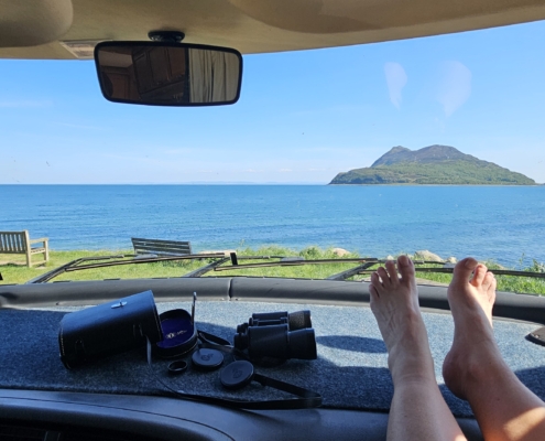 Feet on motorhome dashboard with view of sea and an island through the windscreen