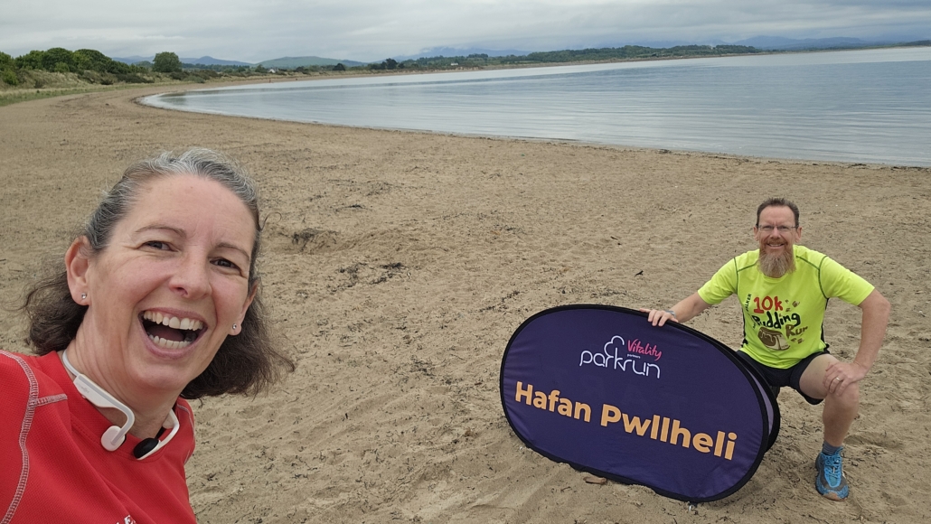 Ju and Jay on a beach next to a Parkrun sign