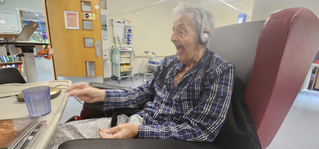Ju's Dad in a chair in his pyjamas listening to a DVD player with headphones and smiling