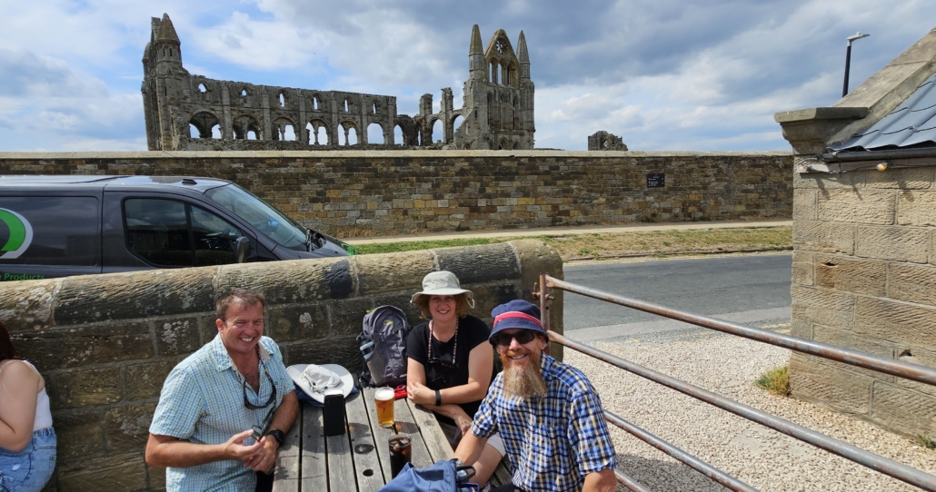 Three people sitting on a picnic bench with Whitby Abbey in the background and beers on the table