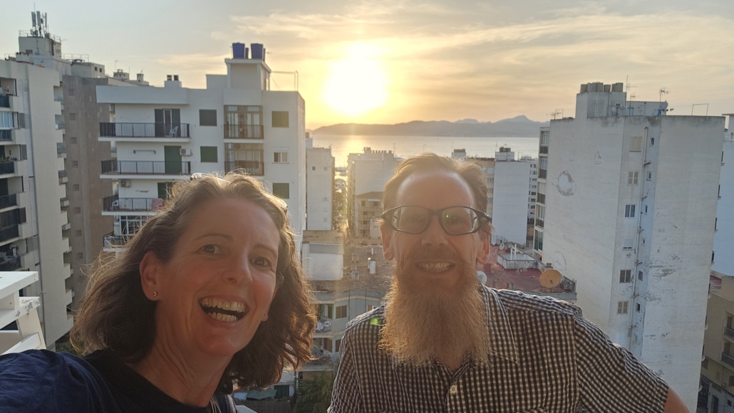 Julie and Jay on a hotel balcony looking over buildings to the sea, hills and sun just above the hills in the sky.