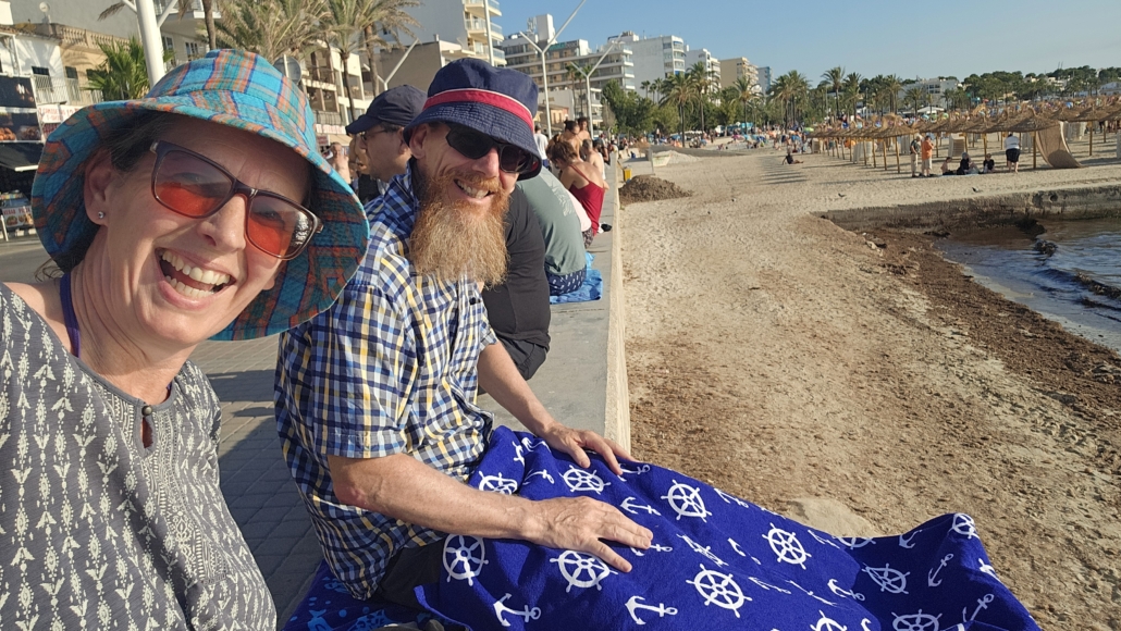 Julie and Jay sitting on a wall by the beach with a towel on Jays legs to protect from the sun