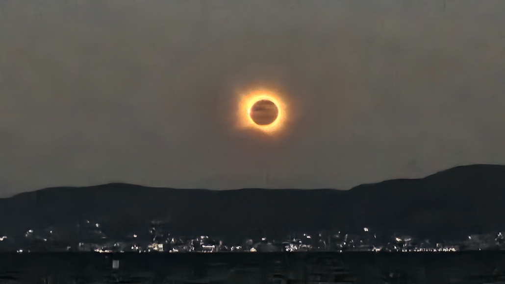 Image of the sun in eclipse above hills and lights on the shore underneath